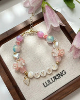 Bracelet with pink beads and 'LOVER' charm on a LULUKING branded card with flowers in the background